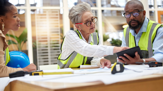 Three people in high-visibility vests sitting around a table with technical plans on it