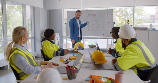 One person in a suit presenting to a group sitting around a table wearing hi-vis vests