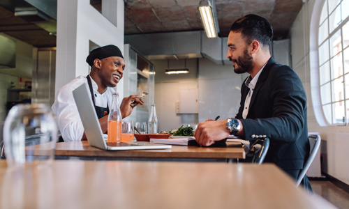 Two people discussing while sitting a table featuring a laptop and some food