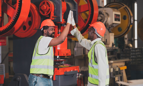 Two employees in hard hats high fiving each other in front of some industrial machinery